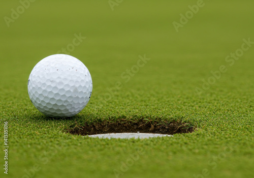 Golf ball perched on edge of hole, green grass in background, blue sky overhead.