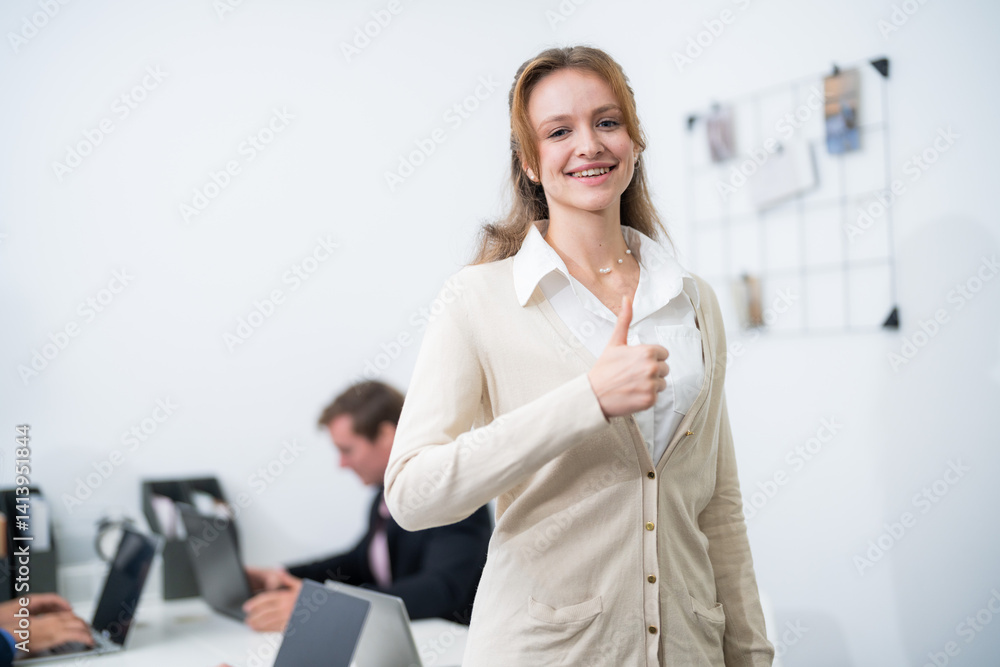 Young woman giving a thumbs up in a modern office while colleagues work at their desks Young woman giving a thumbs up in a modern office while colleagues work at their desks