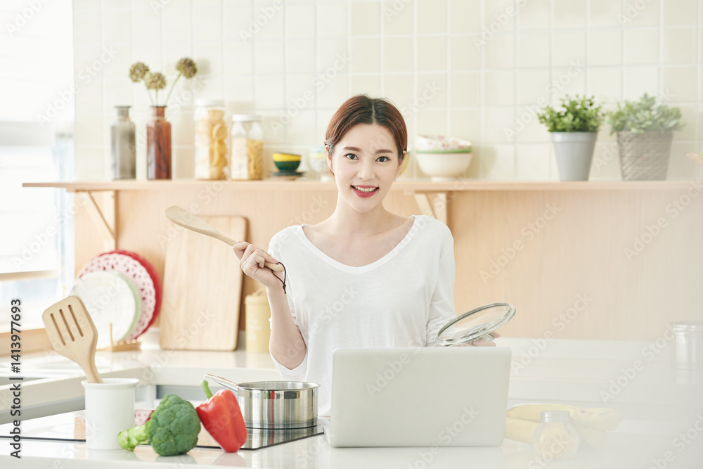 Young Asian Woman Cooking with Laptop in Bright Modern Kitchen, Surrounded by Fresh Vegetables and Kitchen Utensils Young Asian Woman Cooking with Laptop in Bright Modern Kitchen, Surrounded by Fresh Vegetables and Kitchen Utensils