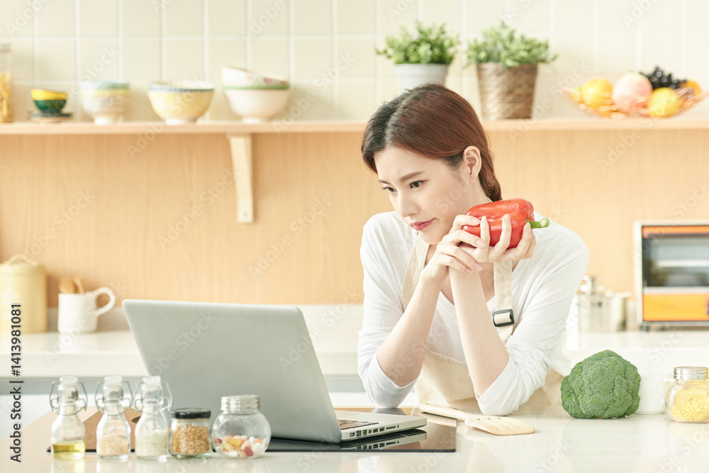 Young Woman Holding Bell Pepper and Using Laptop in Modern Kitchen Young Woman Holding Bell Pepper and Using Laptop in Modern Kitchen