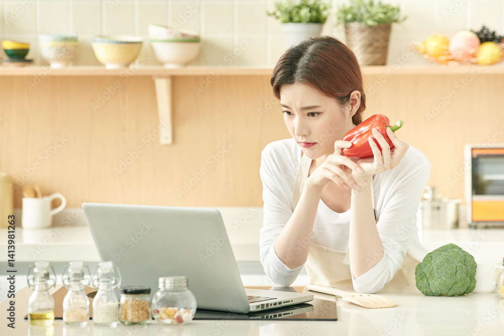 Young Woman Holding Bell Pepper and Using Laptop in Modern Kitchen Young Woman Holding Bell Pepper and Using Laptop in Modern Kitchen