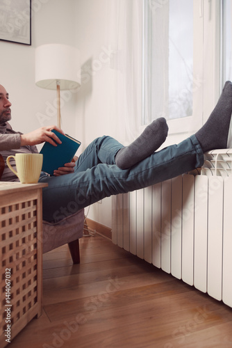 Man's feet with socks on a home heating radiator on a cold winter day outdoors while reading a book and drinking coffee tea from a cup.