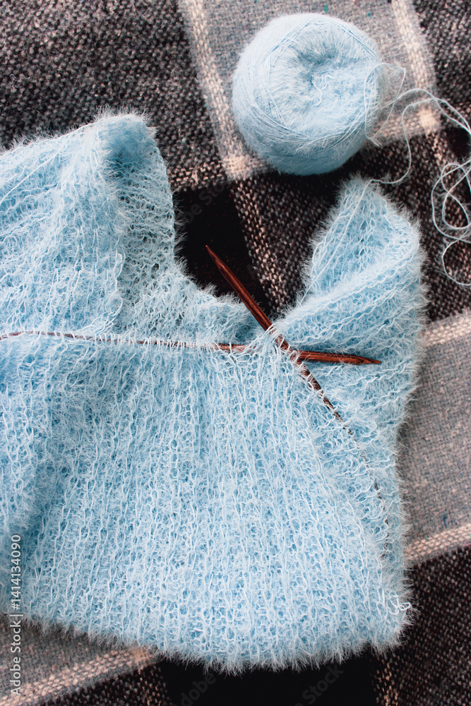 Blue cotton knitting with knitting needles on plaid. Soft blue knitwear with wooden circle needles. Knitting pattern background. Knitting lifestyle. Cozy hobby. Warm knitwear. Craft product concept