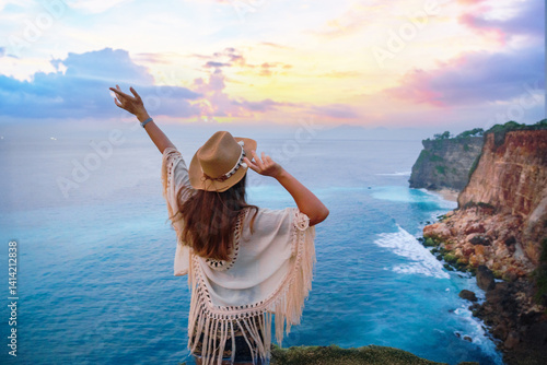 Happy free traveler girl wearing hat with open arms enjoys turquoise stunning ocean view on cliff during traveling at beautiful destination on Bali island, Indonesia