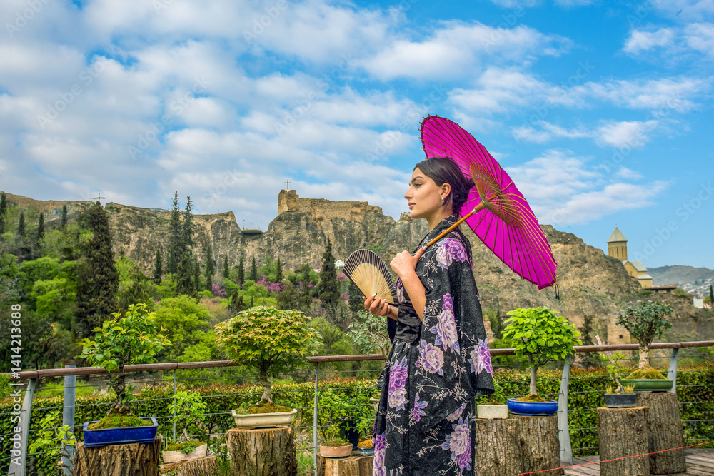 Beautiful young woman in Japanese kimono and close face with fan on holiday of cherry blossom. (sakura ) and hanami. Portrait . . Bonsai trees n wooden stands. Behind Botanical garden and fort Na
