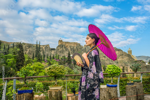 Obraz na płótnie Beautiful young woman in Japanese kimono and close face with fan on holiday of cherry blossom. (sakura ) and hanami. Portrait . . Bonsai trees n wooden stands. Behind Botanical garden and fort Na w salonie