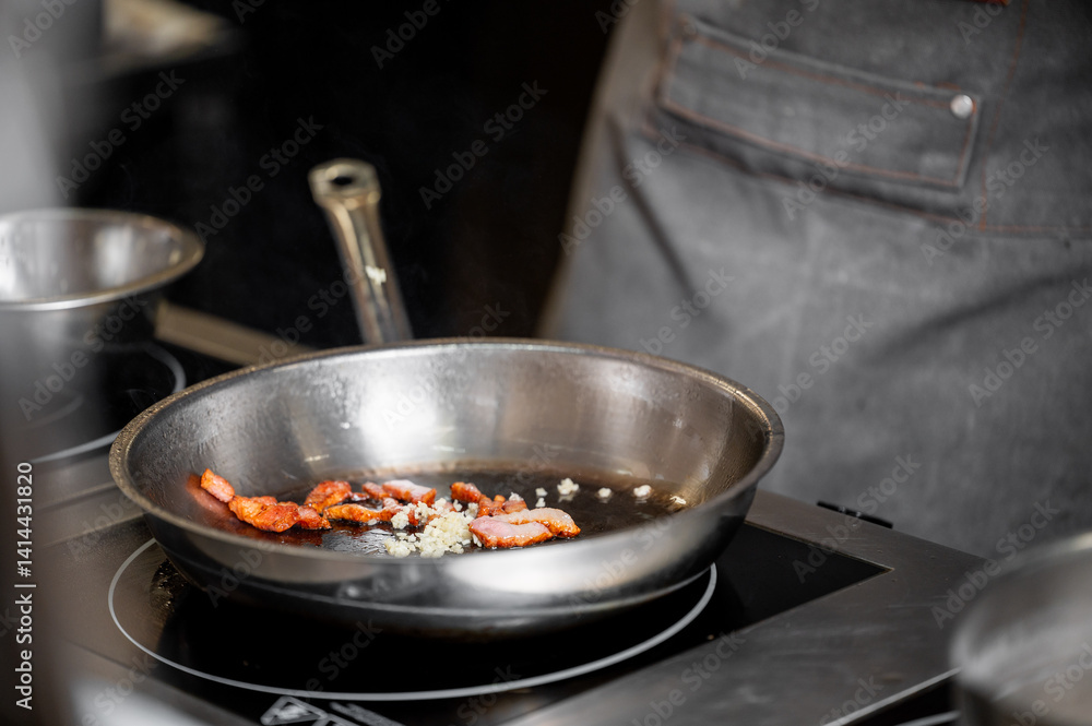 A close-up of sizzling bacon pieces and chopped garlic in a stainless-steel skillet on a stovetop, highlighting a chef's apron in the background in a professional kitchen. A close-up of sizzling bacon pieces and chopped garlic in a stainless-steel skillet on a stovetop, highlighting a chef's apron in the background in a professional kitchen.