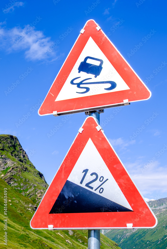 Warning signs alert drivers to curves and steep slopes on mountain road during sunny day in a scenic landscape