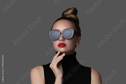Eye wear beauty fashion portrait of a woman in square sunglasses and red lips wearing a black turtleneck. Her hair is styled in a  high bun, giving off a bold and elegant  look on a gray background