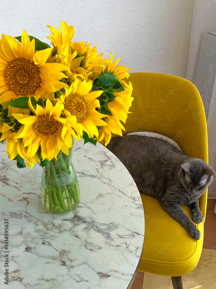 A grey cat is lying on a yellow armchair near a table with a bouquet of sunflowers