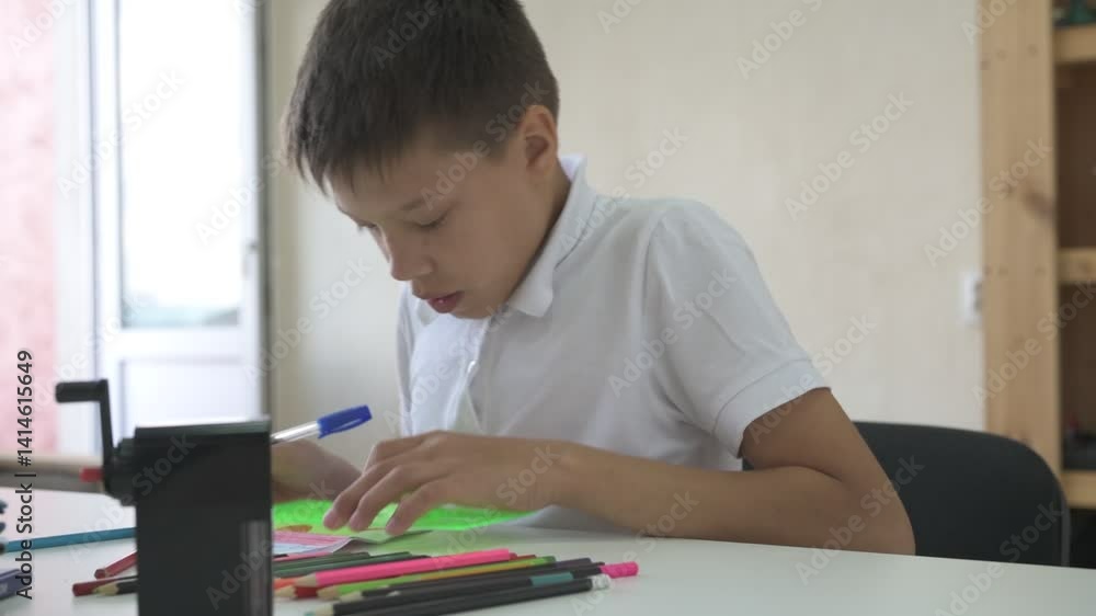 A homeschooled schoolboy does his homework. The boy sits at the table with textbooks and colored pencils A homeschooled schoolboy does his homework. The boy sits at the table with textbooks and colored pencils