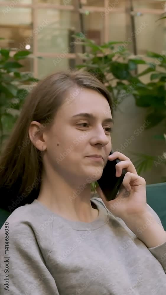 Teenage girl in gray sweater and long dark blonde hair discussing funny story with her friend sitting on sofa in a room surrounded by green plants Teenage girl in gray sweater and long dark blonde hair discussing funny story with her friend sitting on sofa in a room surrounded by green plants