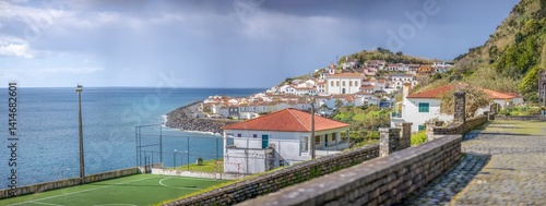 Ribeira Quente, São Miguel, Azoren: Seitlicher Blick entlang der Küste, Fußballplatz und Igreja de São Paulo im Vordergrund, rote Dächer, dramatische Wolken und blaues Meer im Sonnenschein.