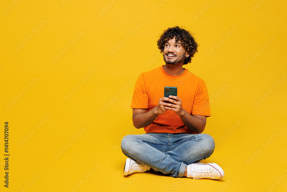 Φωτογραφία Full body young Indian man wear orange t-shirt casual clothes sit hold in hand use mobile cell phone look aside on area mockup isolated on plain yellow background studio portrait