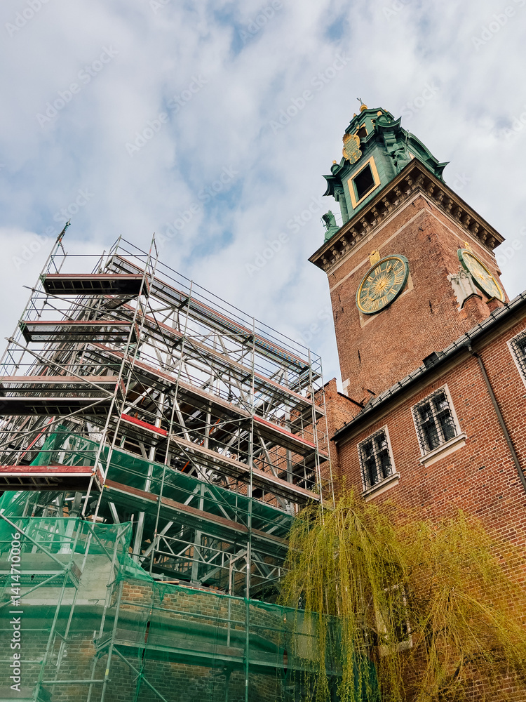 Fototapeta premium Historic Wawel Royal Castle restoration in progress, scaffolding partially covering medieval architecture, landmark clock tower rising against overcast skies in Krakow, Poland. Renovation of Wawel