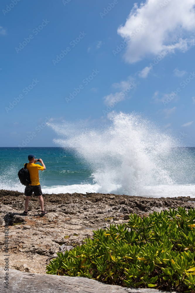 Back View of a Man with Yellow T-Shirt, Baseball Cap and Black Backpack Standing in Front of the Caribbean Ocean Photographing Water Shooting Out from a Blow Hole