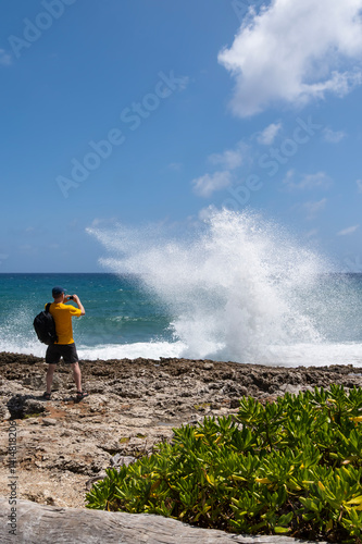 Obraz na płótnie Back View of a Man with Yellow T-Shirt, Baseball Cap and Black Backpack Standing in Front of the Caribbean Ocean Photographing Water Shooting Out from a Blow Hole w salonie