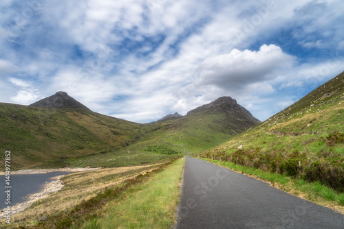 A picturesque and charming view of a winding road that meanders alongside tranquil and peaceful Silent Valley Reservoir, set against lush green Mourn Mountains beneath beautiful sky, Northern Ireland