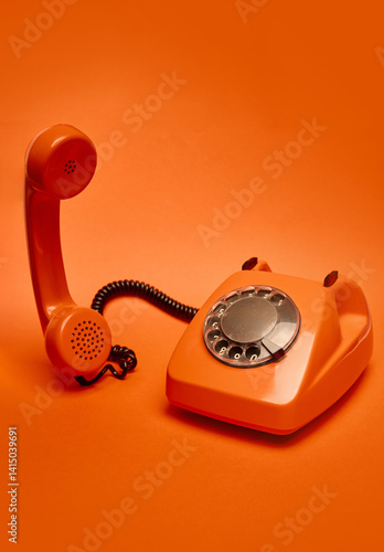 Orange vintage rotary dial telephone with receiver slightly lifted off its base is positioned against monochromatic orange backdrop. Concept of retro, vintage, technology, pop art.