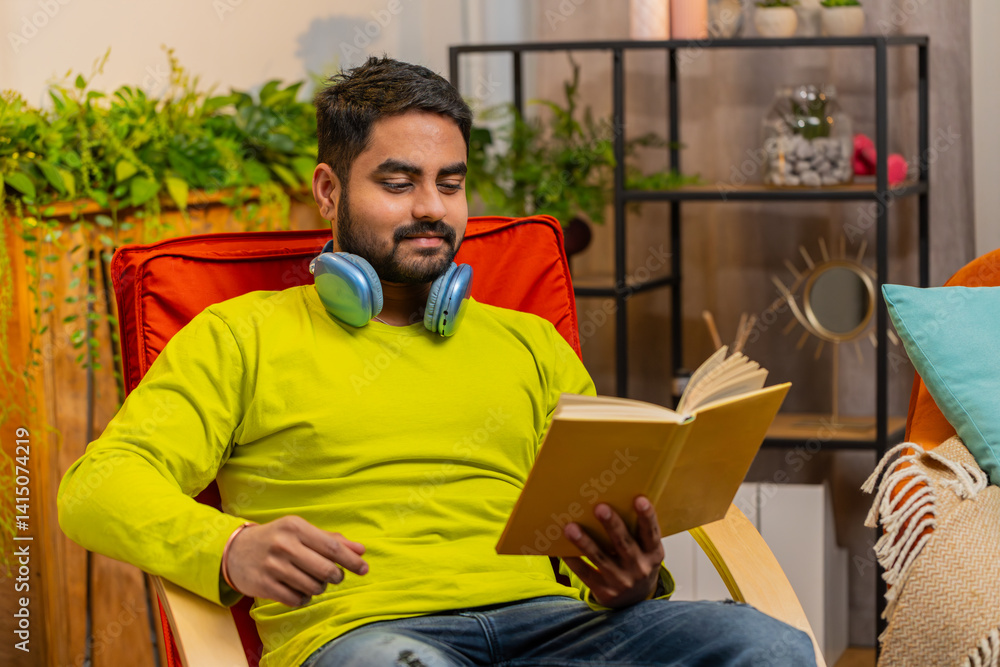Fototapeta premium Indian man reading interesting book, turning pages smiling enjoying literature, taking a rest on comfortable chair. Portrait of peaceful cheerful guy relaxing at home apartment living room on chair
