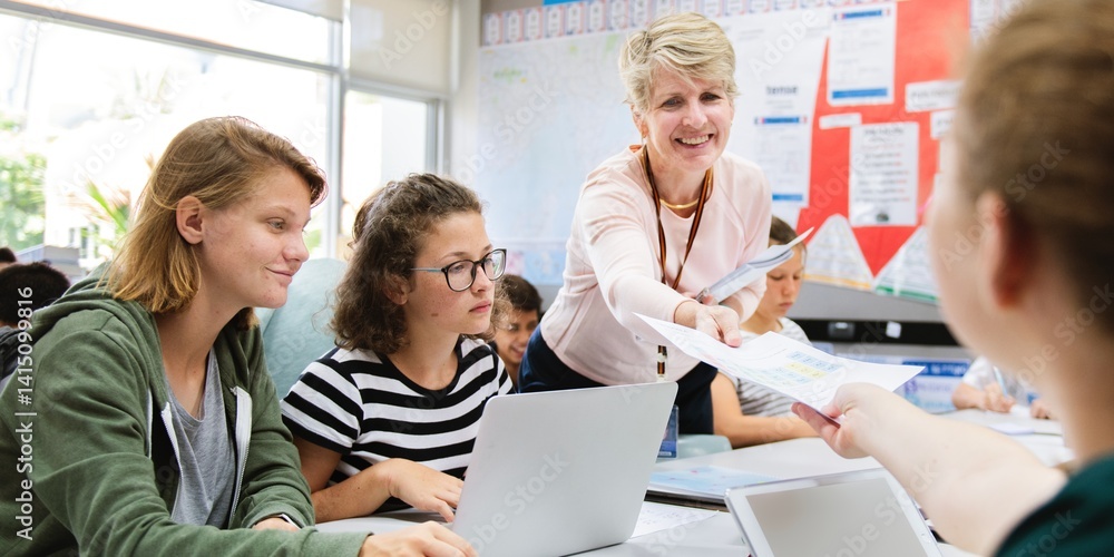 Obraz premium Classroom with diverse students and teacher, work on school group project. Diverse students work on laptop in classroom. Teacher engages with students, group project collaboration in school classroom