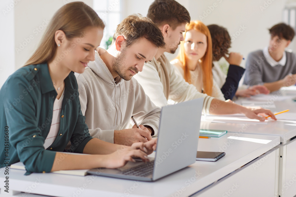 Group of young confident college or university students sitting at the same desk in the classroom during a lesson doing educational project on laptop. Education and knowledge concept. Group of young confident college or university students sitting at the same desk in the classroom during a lesson doing educational project on laptop. Education and knowledge concept.