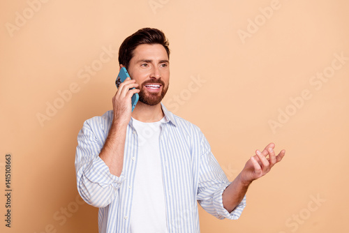 Young man in a striped shirt talking on the phone against a neutral beige background, gesturing positively with expressive smile