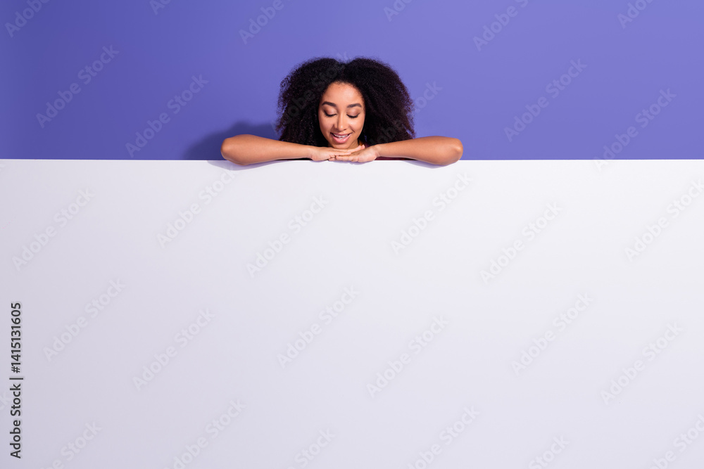 Portrait of a stylish young woman with curly hair peeking over a blank board against a purple background Portrait of a stylish young woman with curly hair peeking over a blank board against a purple background