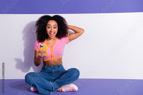 Young woman with curly hair holding a phone, sitting cross-legged on a purple and white background
