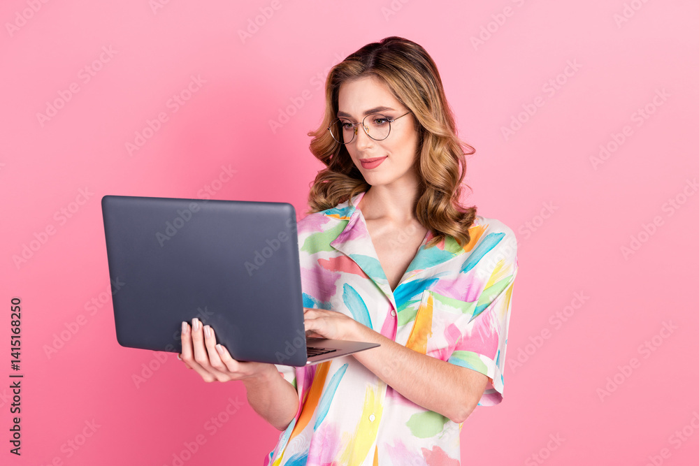 Young woman with stylish shirt and glasses using a laptop against vibrant pink background with a confident and focused demeanor Young woman with stylish shirt and glasses using a laptop against vibrant pink background with a confident and focused demeanor