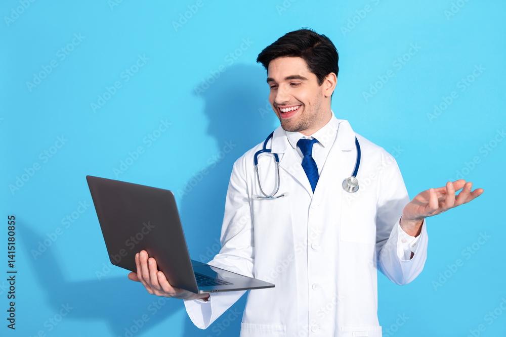 Smiling young doctor holding a laptop against a bright blue background wearing a white coat and stethoscope Smiling young doctor holding a laptop against a bright blue background wearing a white coat and stethoscope