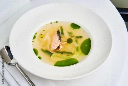 A bowl of fragrant fish soup is elegantly presented, featuring tender pieces of fish nestled in a clear broth with vivid green herbs. This dish invites warmth and comfort, perfect for a gourmet meal