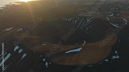 Aerial view of a volcanic crater with streaks of snow on dark ash slopes, captured in Iceland during golden hour light.