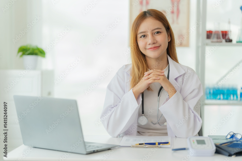 Smiling Asian female doctor in lab coat and stethoscope sitting next to online consultation table in doctor's office Smiling Asian female doctor in lab coat and stethoscope sitting next to online consultation table in doctor's office
