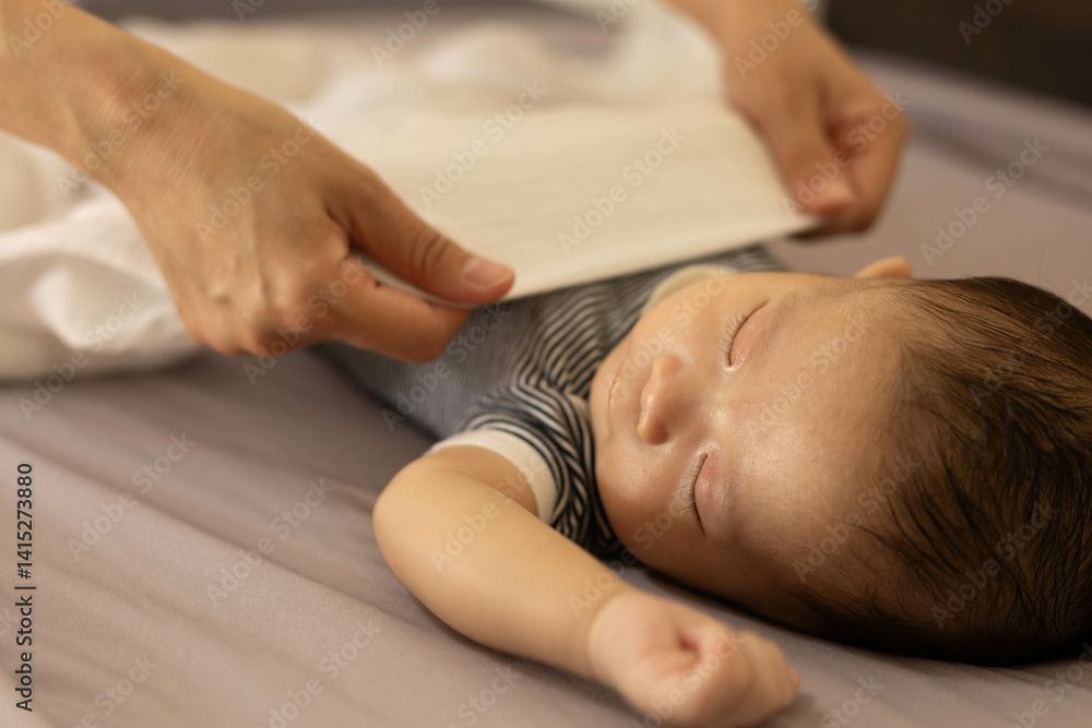 A baby is sleeping on a bed with a blanket being pulled over him. The baby is wearing a blue and white striped shirt A baby is sleeping on a bed with a blanket being pulled over him. The baby is wearing a blue and white striped shirt