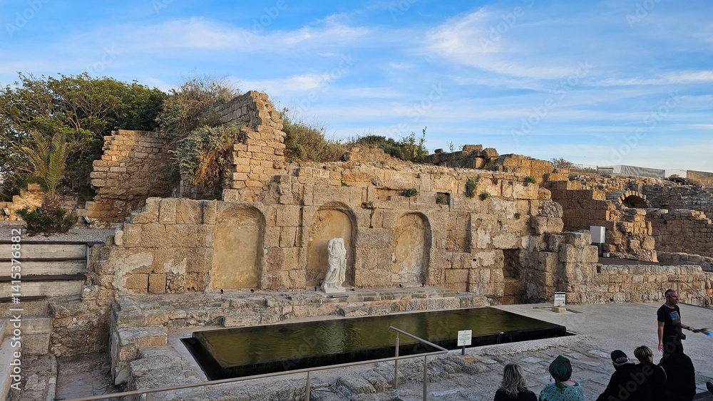 Obraz premium Ancient Roman bath ruins at Caesarea Maritima, Israel, with a reflecting pool and visitors exploring the site.