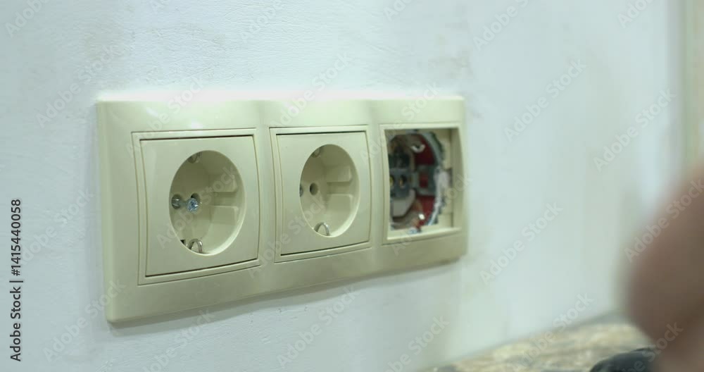 A worker mounts a double socket on a white wall and screws it with an electric drill.