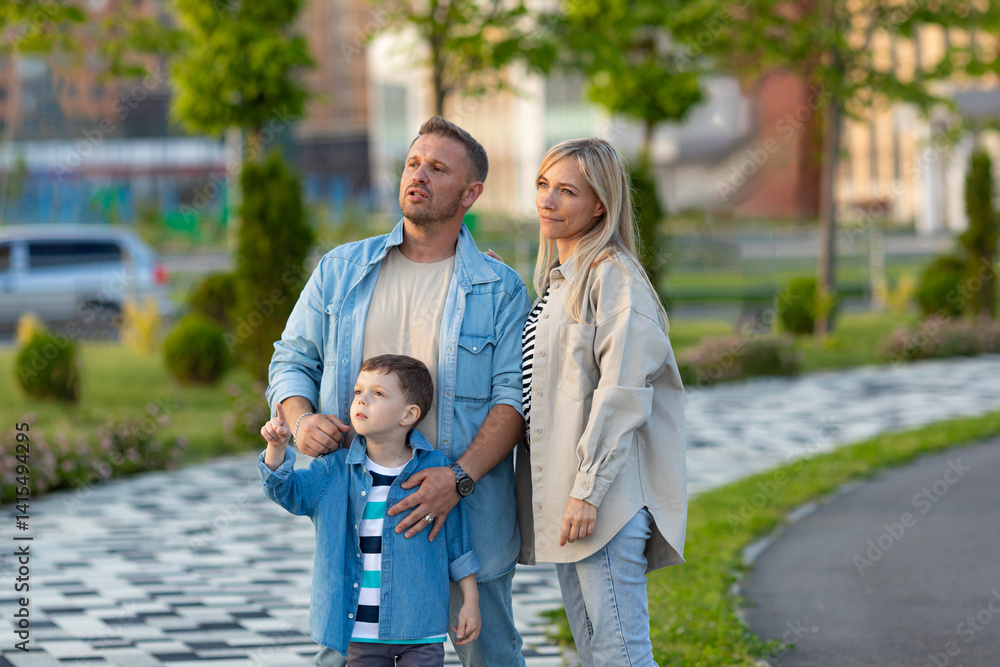 Happy family concept. Dad, mom and little boy son spend time together, walking in city park on summer vacation. Parents hugging his son and all they looking far away. Lifestyle at city public park