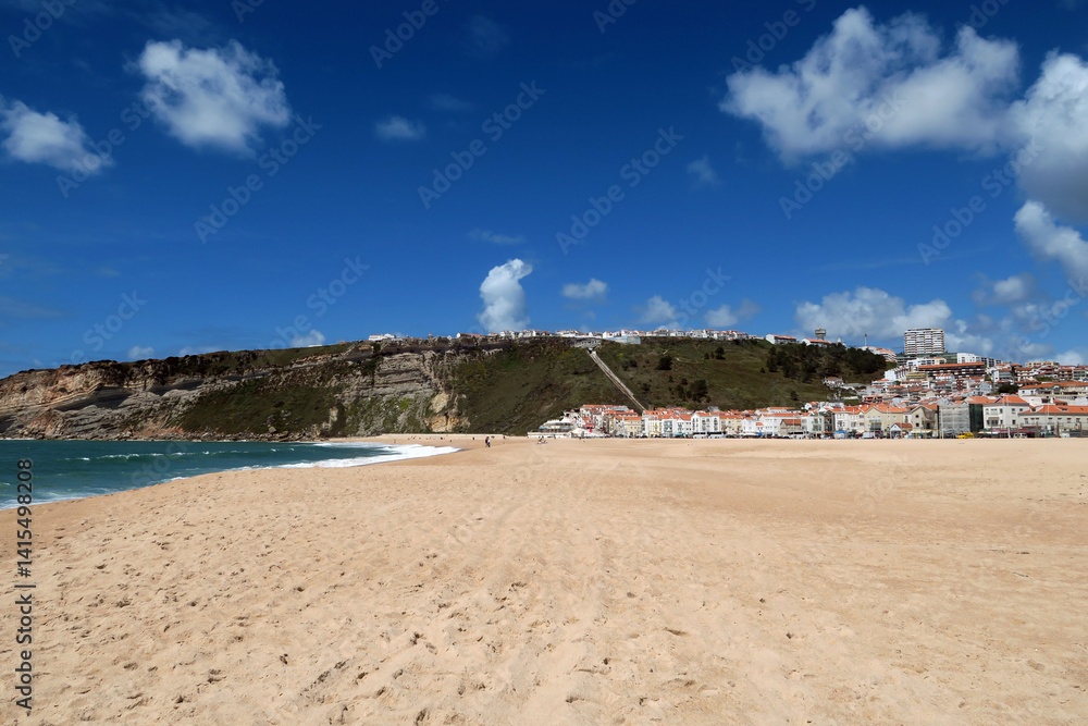 A sunny beach scene with golden sand, gentle waves and a rocky cliff topped with houses under a vibrant blue sky in Nazaré, Portugal. A sunny beach scene with golden sand, gentle waves and a rocky cliff topped with houses under a vibrant blue sky in Nazaré, Portugal.