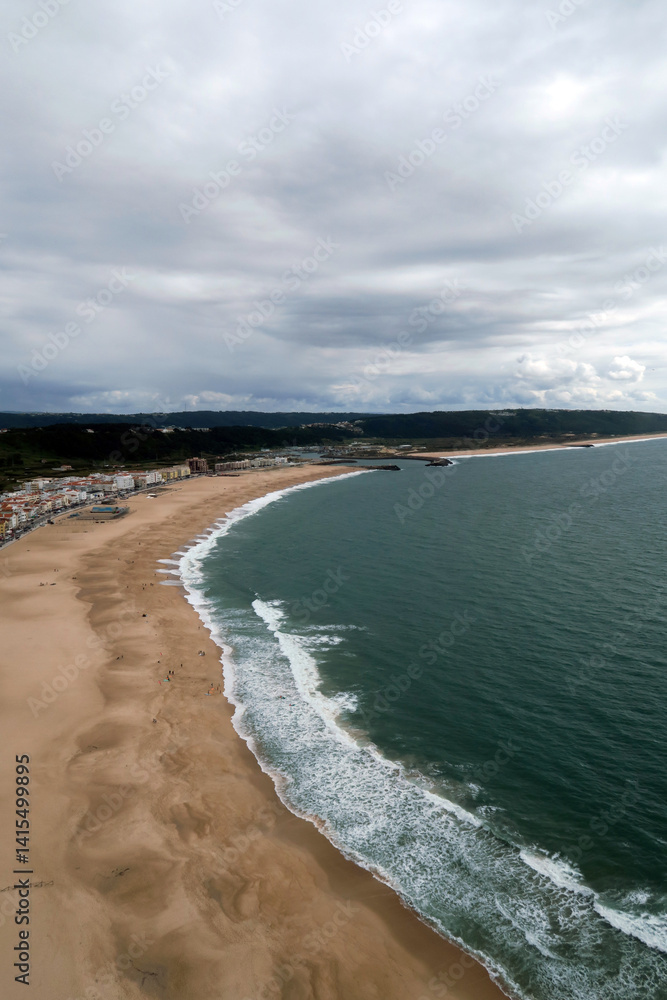 A sunny beach scene with golden sand, gentle waves and a rocky cliff topped with houses under a vibrant blue sky in Nazaré, Portugal. A sunny beach scene with golden sand, gentle waves and a rocky cliff topped with houses under a vibrant blue sky in Nazaré, Portugal.