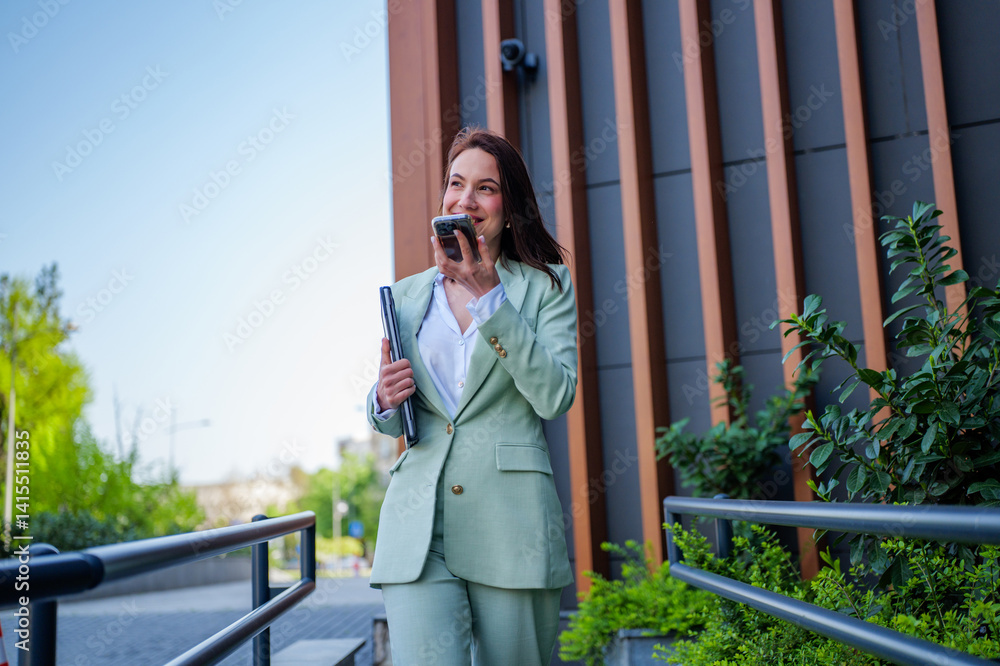 Fototapeta premium Smiling businesswoman holding a laptop and chatting on a mobile phone while leaving the office, enjoying a successful day at work as she walks through the vibrant city streets