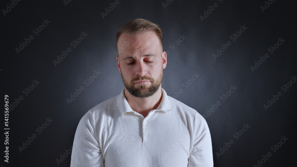 A man in a white shirt stands with eyes closed, conveying a sense of contemplation or exhaustion. The dark background accentuates his introspective mood. A man in a white shirt stands with eyes closed, conveying a sense of contemplation or exhaustion. The dark background accentuates his introspective mood.