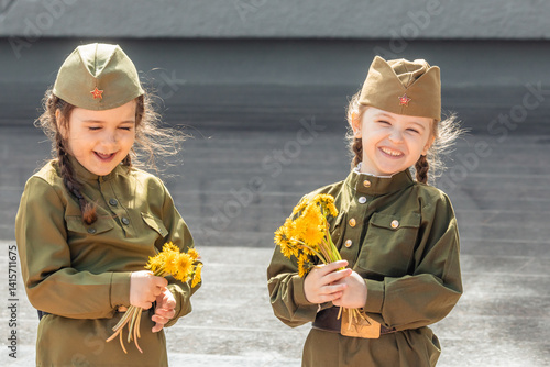 Obraz na płótnie Patriotic education of children. Little girls with long braids in a military uniform, military cap, hold flowers in hands against the background of the memorial. Photo for the Victory Day on May 9 w salonie