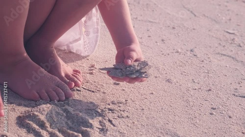 Child crouched on a sandy beach, holding sand in one hand and sifting through it with the other, barefoot. Focus on hands and feet, depicting a playful beach moment