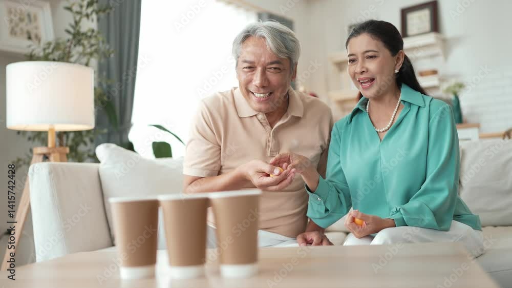 Happy elderly couple enjoying a ball toss game together at home, practicing coordination and cognitive skills for healthy aging. Grandfather and grandmother throwing ping pong in the bowl. Myrmidon. Happy elderly couple enjoying a ball toss game together at home, practicing coordination and cognitive skills for healthy aging. Grandfather and grandmother throwing ping pong in the bowl. Myrmidon.