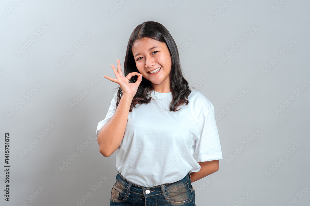 Fototapeta premium Happy young Asian woman showing an OK hand sign while smiling confidently at the camera. Studio portrait in casual outfit, conveying approval, satisfaction, and friendliness.