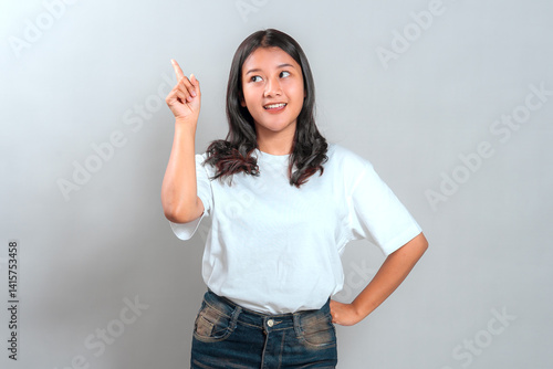 Photography Cheerful young Asian woman with bright facial expression, smiling and pointing upward with one finger, showing she just got an idea or inspiration, standing against plain background