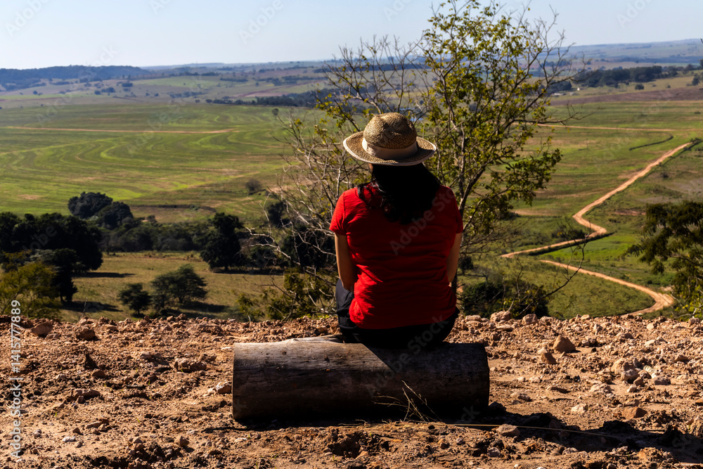 Woman sitting on a wooden stump looks out over the landscape of rural roads and farmland from a mountain range in Brazil