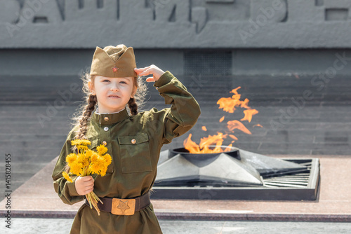 Obraz na płótnie Patriotic education of children. Little girl with long braids in a military uniform, military cap, holds flowers in hands against the background of the memorial. Photo for the Victory Day on May 9 w salonie