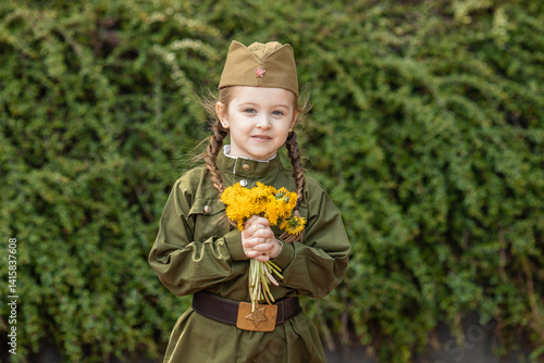 Obraz na płótnie Patriotic education of children. Little girl with long braids in a military uniform, military cap, holds flowers in hands against the green plants background. Photo for the Victory Day on May 9 w salonie